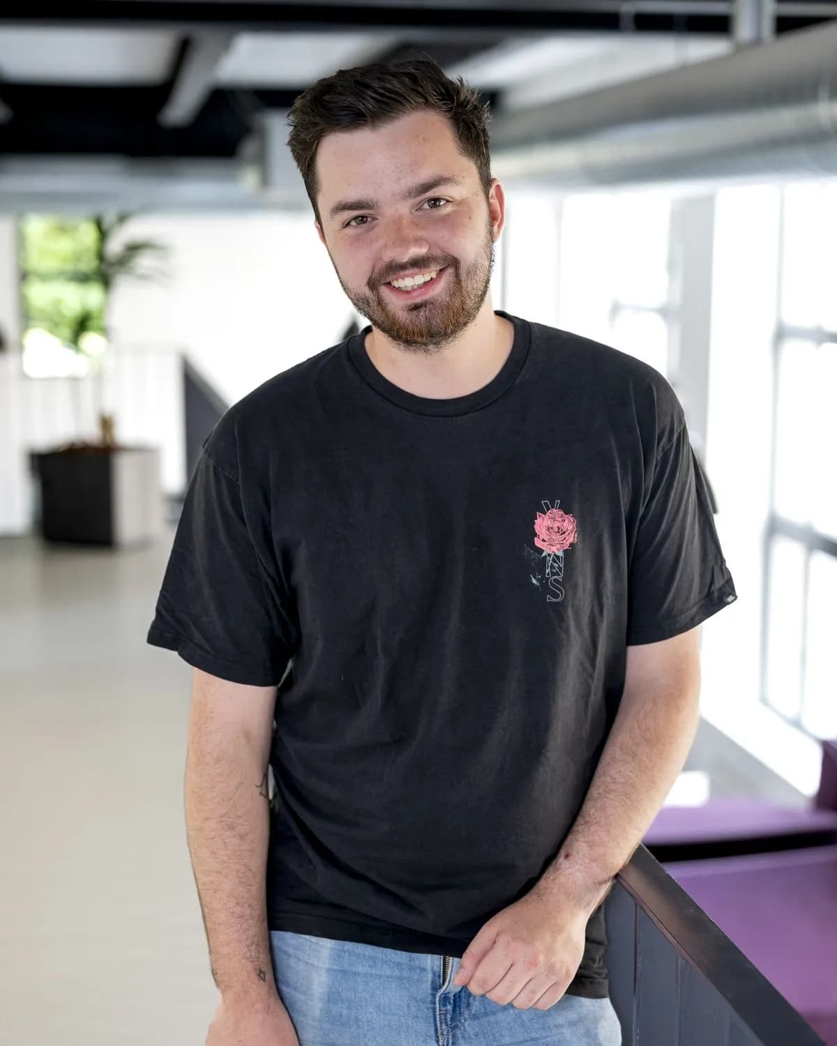 Noel lehnt lächelnd an einem Geländer in einem hellen Büro.